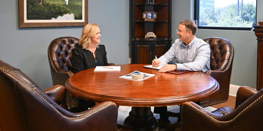 Two people, a woman and a man, sit across from each other at a round wooden table in an office, engaged in conversation. The woman has blonde hair and wears a black blazer, while the man has short, gr.